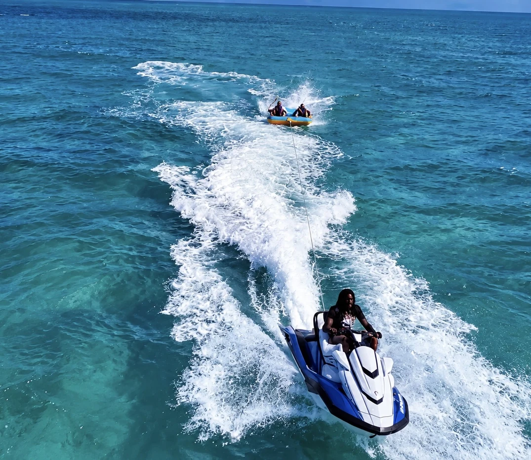 Aerial view of a jet ski and inflatable riders leaving a white wake through turquoise water.