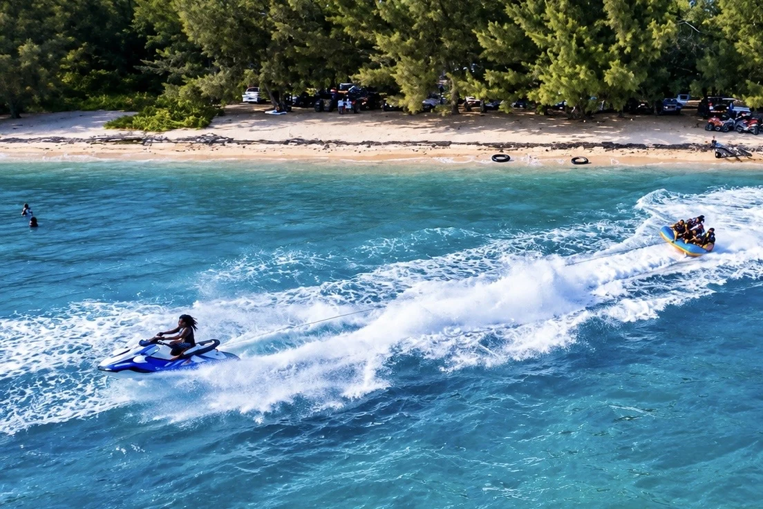 A jet ski towing inflatable riders beside a tree-lined beach, viewed from above.